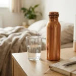 A featured photograph (1024x1024 px) focused sharply on a used, slightly oxidized pure copper bottle and a glass of water resting on a textured wooden bedside table. A small, patterned notebook and a pen are nearby, suggesting a personal morning wellness ritual. The background is softly blurred with subtle, cozy bedroom clutter, emphasizing a natural 'trial and error' feel. Soft ambient daylight.